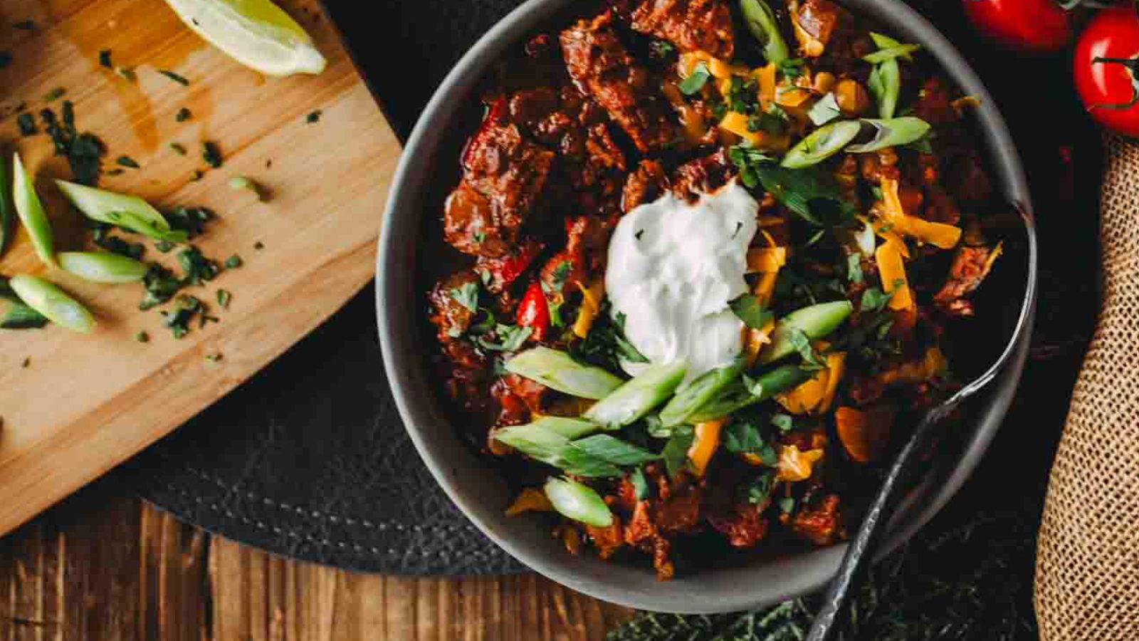 A bowl of chili topped with sour cream, sliced green onions, and cilantro, with a wooden board of lime wedges and chopped herbs nearby. A spoon rests in the bowl.