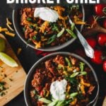 Pinterest interest for Smoked brisket chili; Two bowls of smoked brisket chili topped with sour cream, cheese, and green onions on a wooden table, surrounded by tomatoes, lime, and parsley.