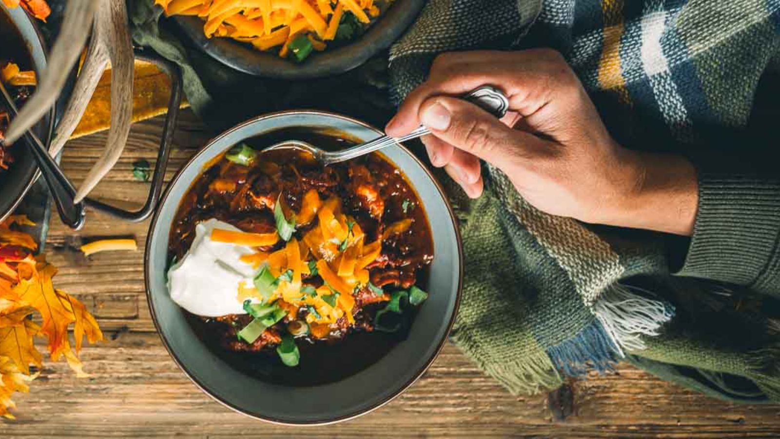 A person holds a spoon over a bowl of chili, crafted from cherished chili recipes, topped with sour cream, shredded cheese, and green onions, all set on a wooden table with a plaid fabric nearby.