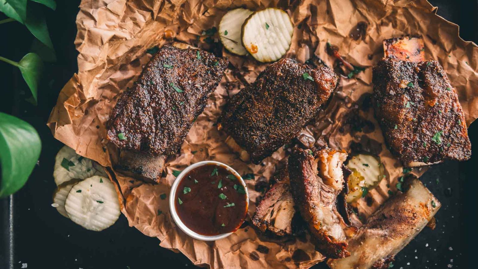 Grilled ribs on brown paper with garnish, sliced pickles, and a small cup of sauce on the side.