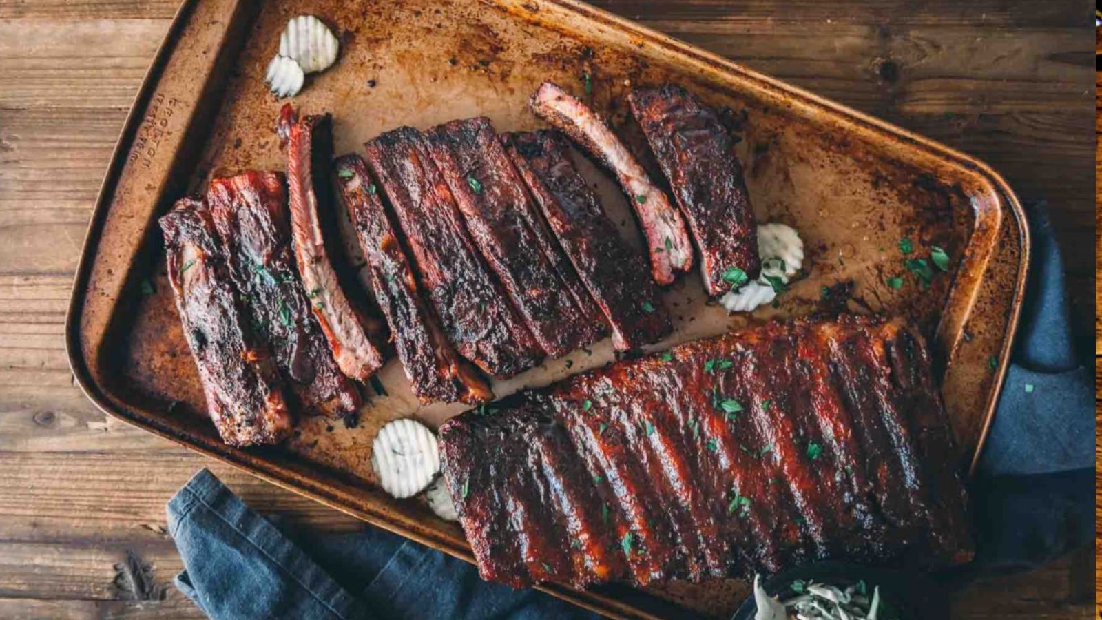 A tray of barbecued ribs with visible grill marks, garnished with herbs. Wavy potato chips are placed next to the ribs.