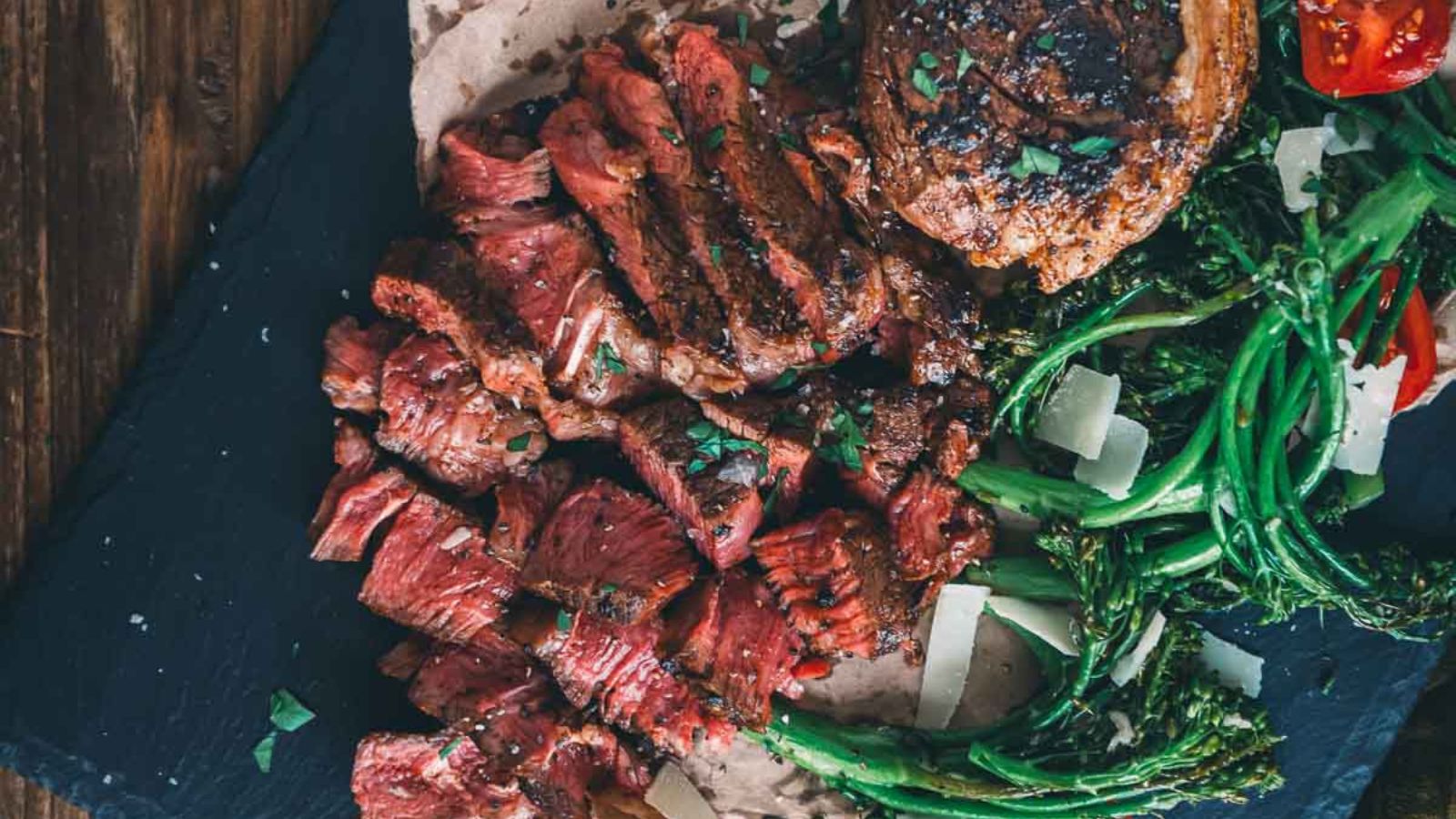 Sliced sous vide chuck steak with herbs, broccolini, and cherry tomatoes served on a dark slate surface.