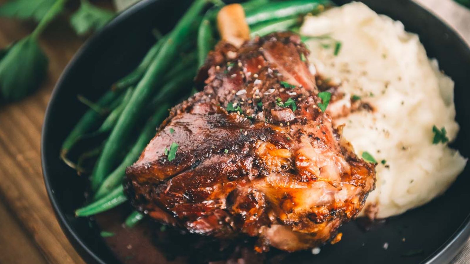 A plate of roasted lamb shank with green beans and mashed potatoes garnished with herbs.