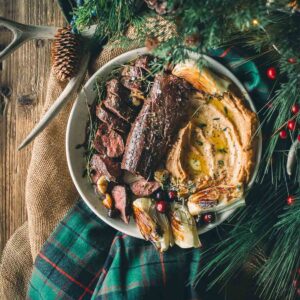 A plate with sliced sous vide venison tenderloin, pureed root veggies, roasted fennel, and herbs, surrounded by festive greenery and a plaid cloth on a wooden table.