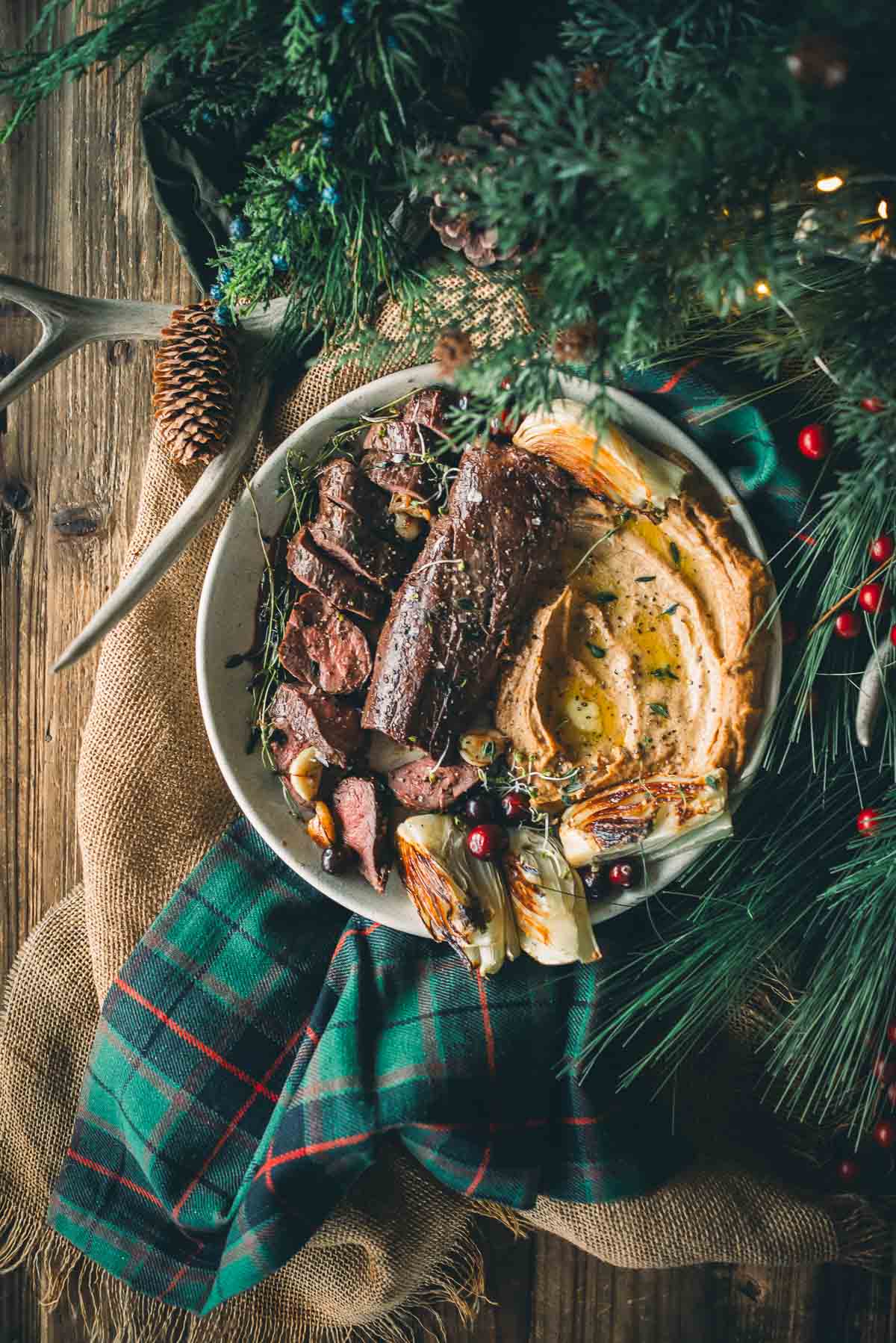 A plate with sliced sous vide venison tenderloin, pureed root veggies, roasted fennel, and herbs, surrounded by festive greenery and a plaid cloth on a wooden table.