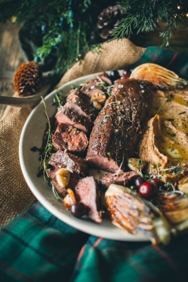 Plate of sliced sous vide venison tenderloinf with herbs, roasted fennel, and cranberries, accompanied by mashed root veg and garnished with thyme sprigs.