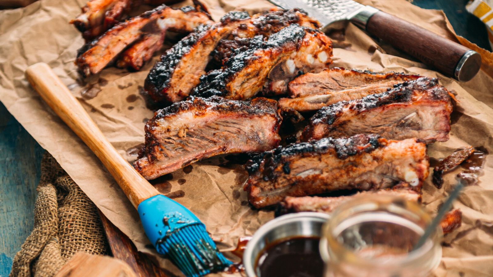 Barbecued ribs on brown paper with a brush, sauce jar, and knife, arranged on a rustic tabletop.