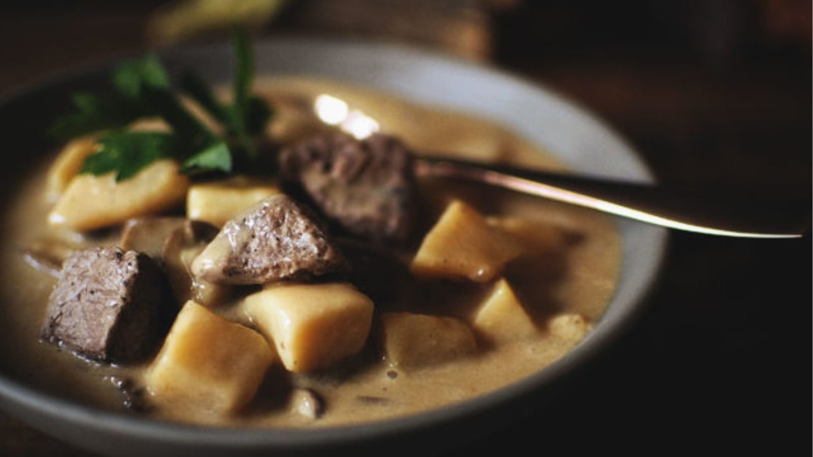 A bowl of beef stew with chunks of beef, potatoes, and a creamy sauce, garnished with parsley. A spoon rests on the edge of the bowl.