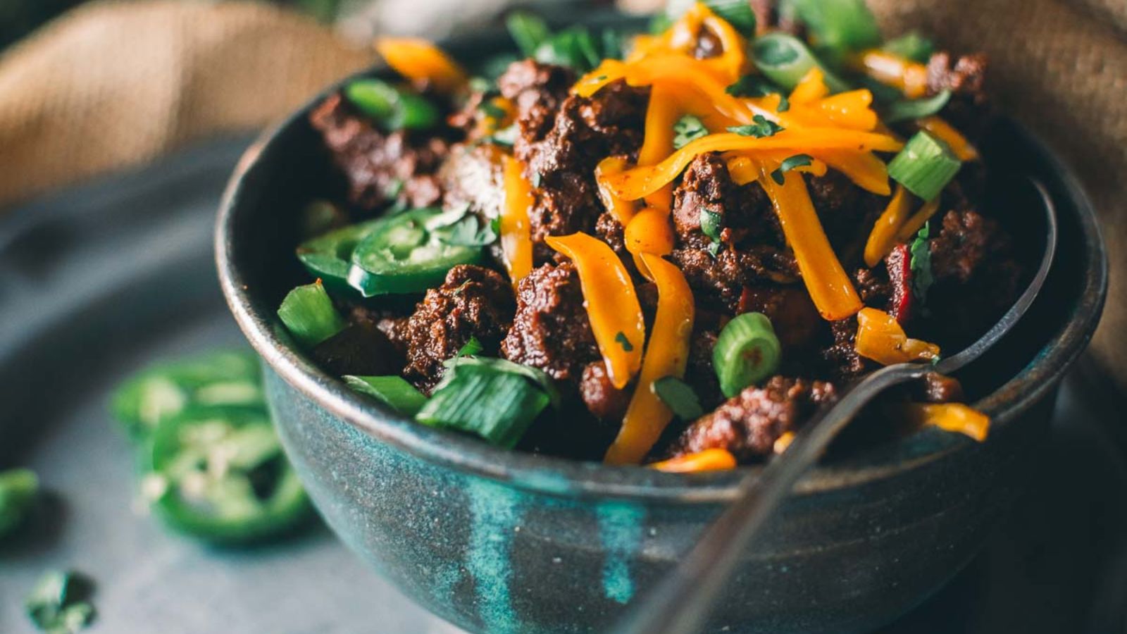 Bowl of chili topped with shredded cheese, sliced jalapeños, and chopped green onions. A spoon rests inside the bowl.