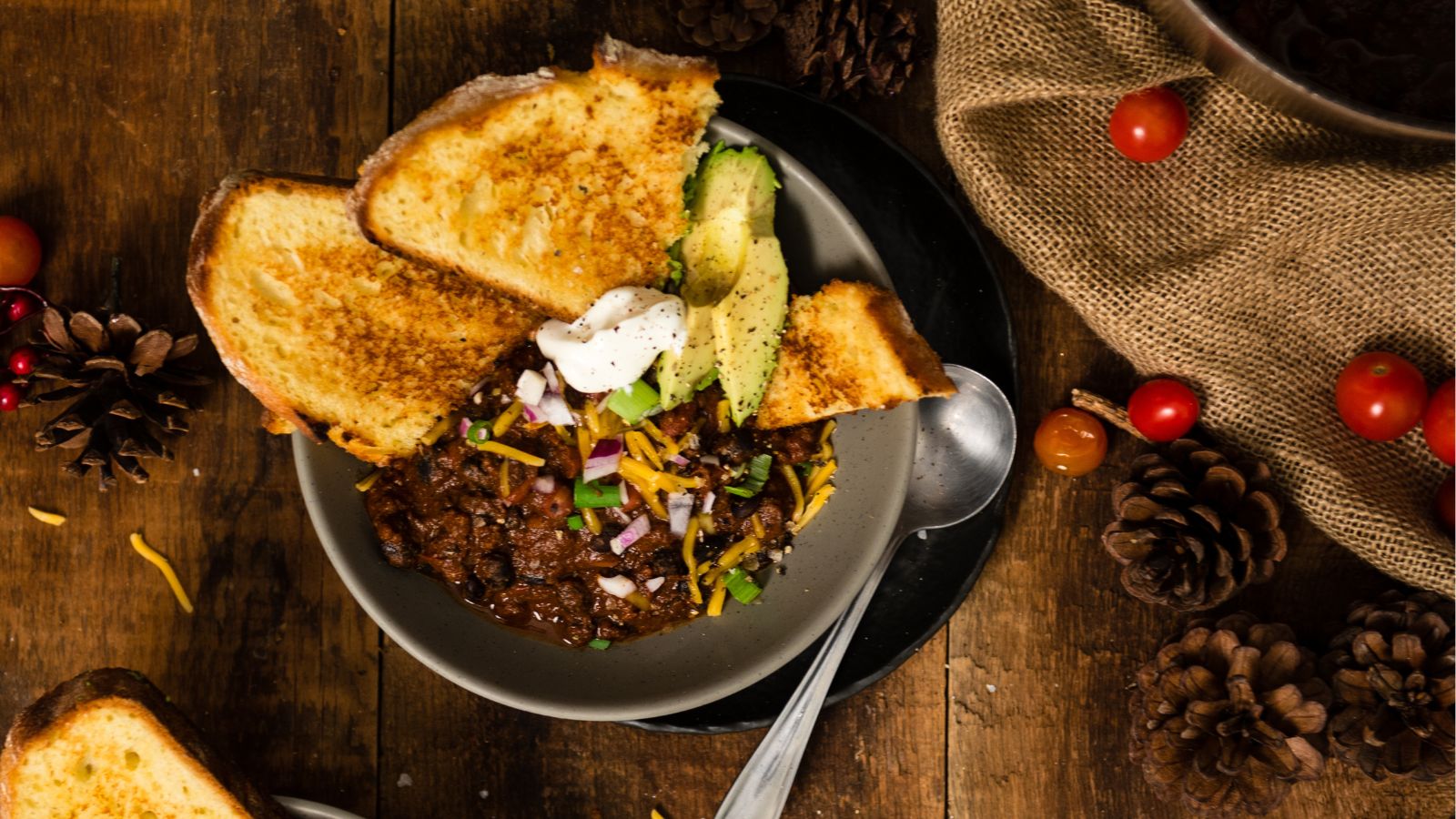 A bowl of chili topped with cheese, red onions, avocado, and sour cream, served with toasted bread.