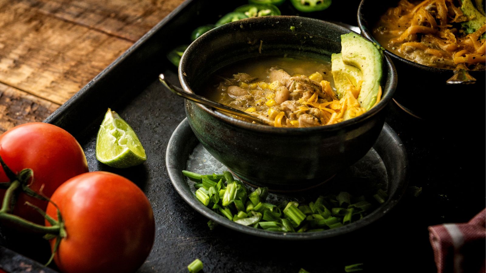 A bowl of soup with meat, avocado, and green onions on a tray. Nearby are sliced jalapeños, a wedge of lime, whole tomatoes, and a dark wooden surface.