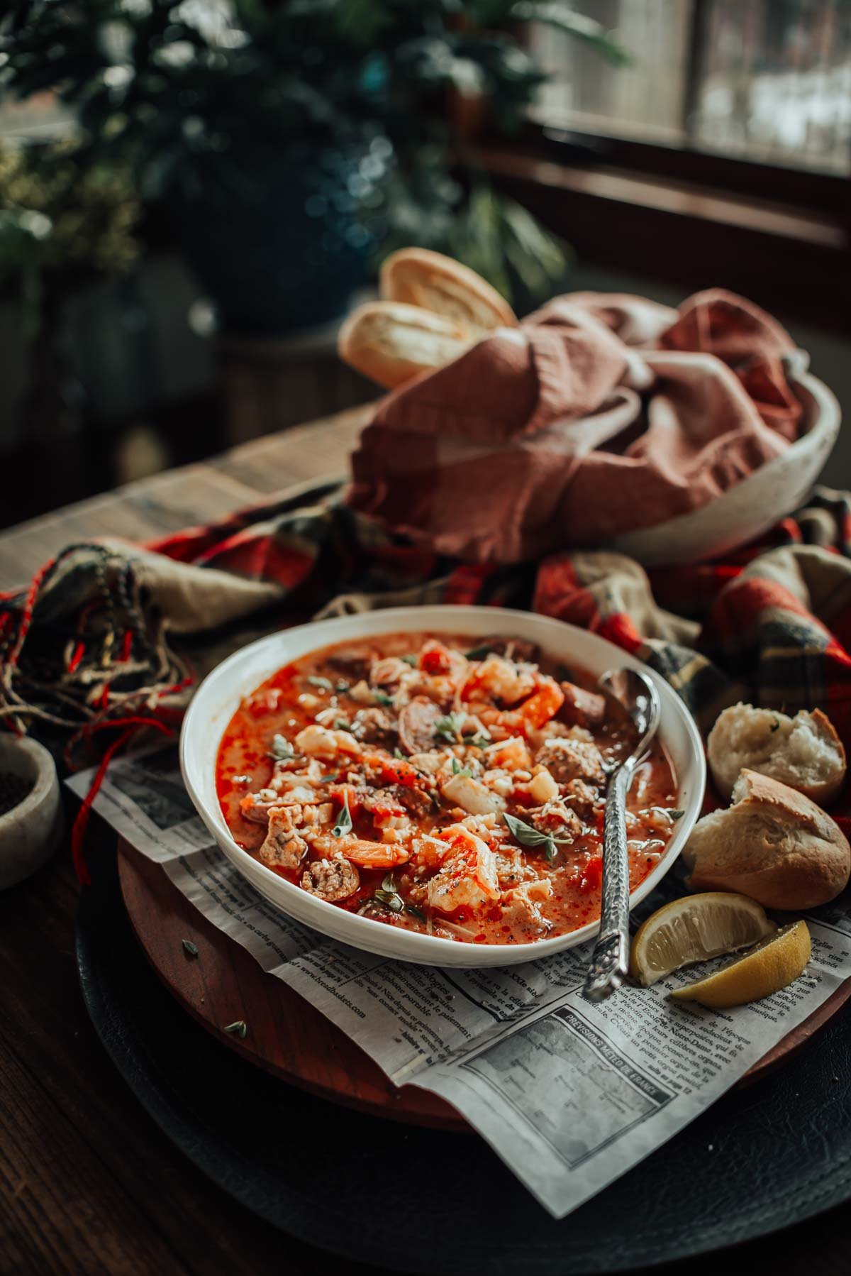 A bowl of hearty seafood stew with vegetables and meat sits on a wooden table, surrounded by bread, a lemon wedge, and a cloth.