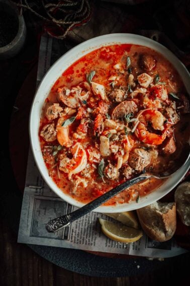 A bowl of seafood stew with shrimp, sausage, and herbs in a red broth, accompanied by bread slices and a lemon wedge on the side.