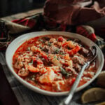 A bowl of seafood stew with shrimp, spices, and herbs on a wooden table, accompanied by a spoon and lemon wedges. A patterned cloth is in the background.