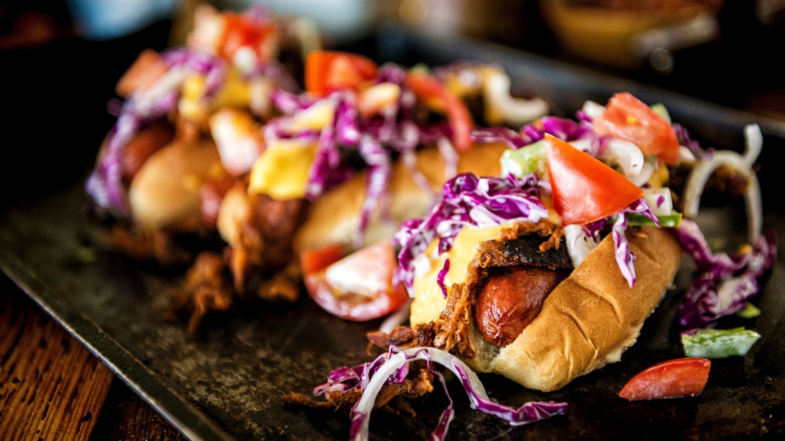 Three hot dogs topped with coleslaw, diced tomatoes, and cheese on a tray.