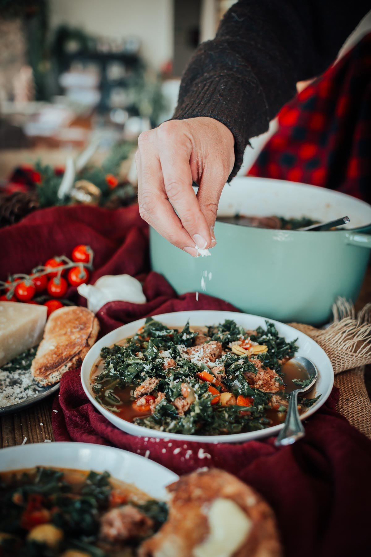 A hand sprinkles cheese over a bowl of soup with kale and sausage with more cheese, a pot, cherry tomatoes, and bread are on the table.