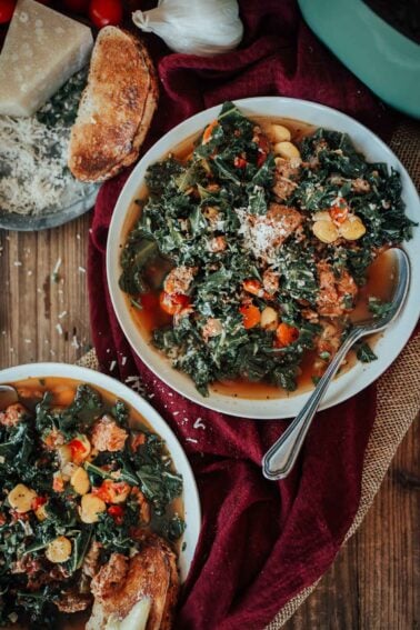 Bowls of soup with sausage and kale, beans, and vegetables garnished with grated cheese; a spoon rests in one bowl. Toasted bread and cheese are on the side.