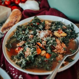 A bowl of soup with kale, sausage, carrots, and shredded cheese. A spoon rests in the bowl on a burgundy cloth, with cherry tomatoes and a pot in the background.