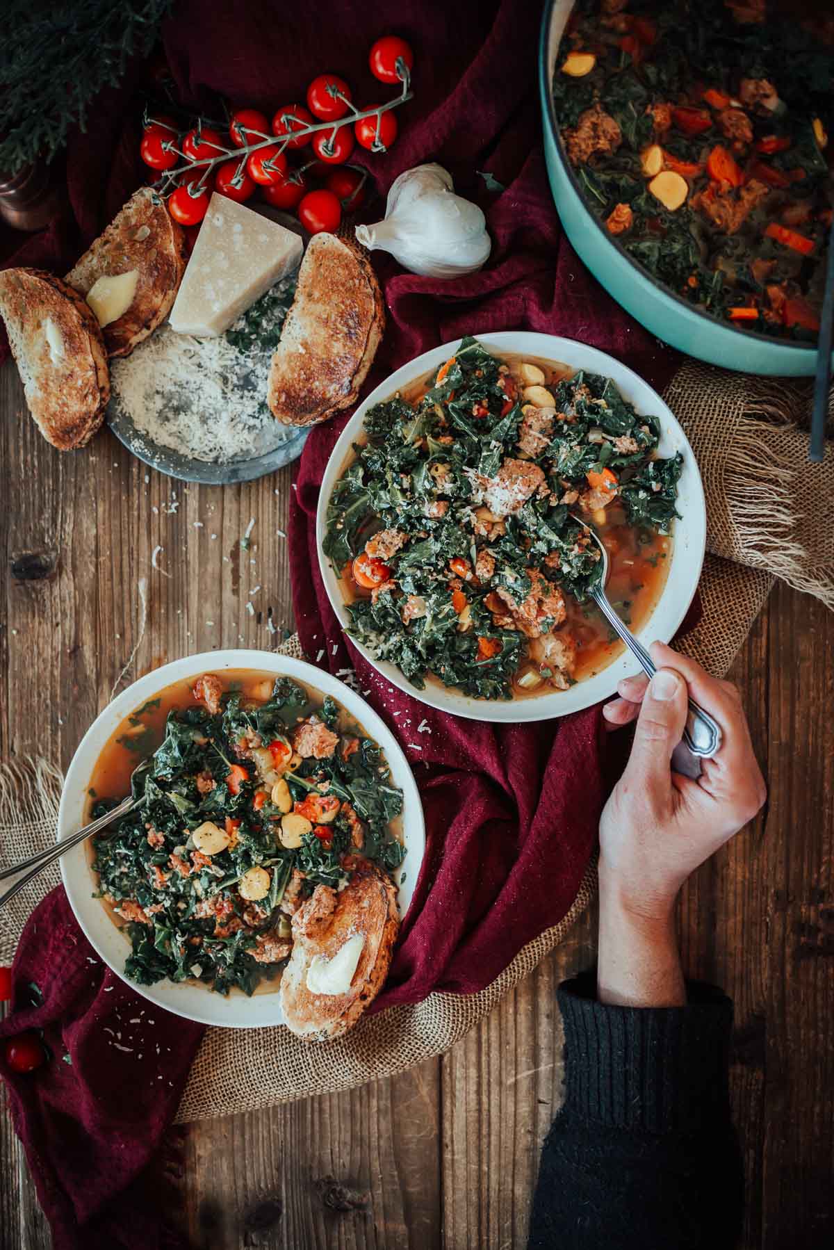 Two bowls of hearty sausage and kale soup with a hand holding a spoon digging in, on a wooden table, surrounded by bread, cheese, garlic, and red fabric.