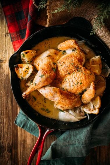 Crispy skined chicken in a red skillet with lemon garlic sauce, onions, and herbs, set on a wooden table with a green cloth and plaid fabric.