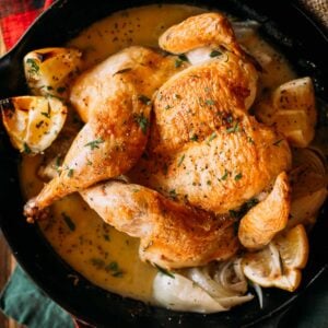 Roasted whole chicken in a cast iron skillet with lemon wedges, herbs, and onions and luxurious sauce.