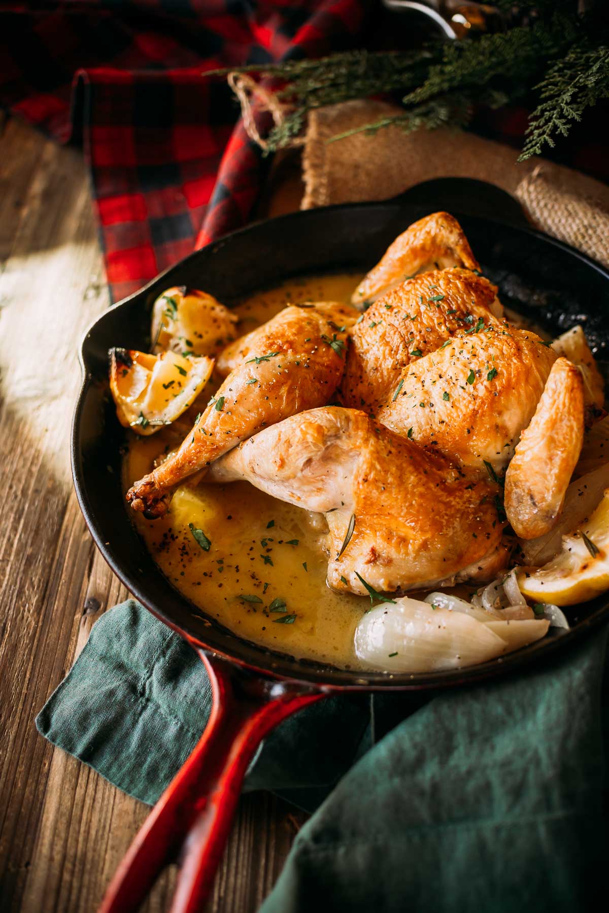 Baked spatchcocked chicken with herbs and lemon sauce in a cast iron skillet, placed on a green cloth with a red checkered fabric in the background.