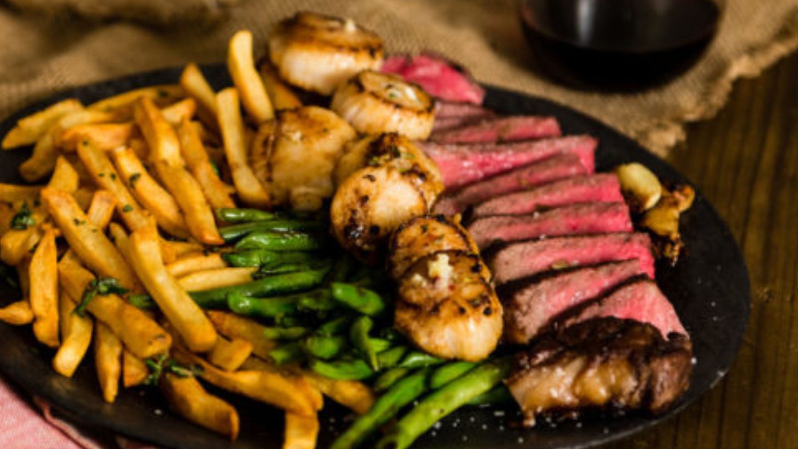 A plate featuring a delectable surf and turf feast perfect for Valentine's Day, with sliced steak, grilled scallops, green beans, and crispy fries.