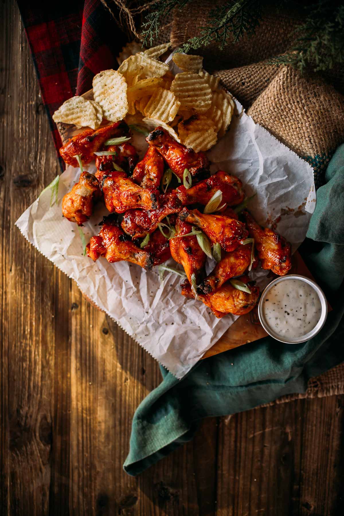 honey sriracha wings topped with green onions on parchment paper, served with potato chips and a small container of dipping sauce, placed on a wooden surface.