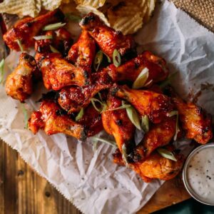 A platter of honey sriracha wings garnished with sliced green onions, served with crinkle-cut chips and a small bowl of dipping sauce on the side.