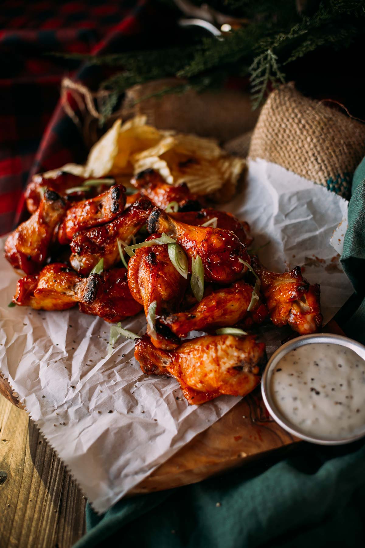 A serving of spicy honey sriracha wings garnished with green onions, placed on crumpled parchment paper, with a side of dipping sauce and potato chips in the background.