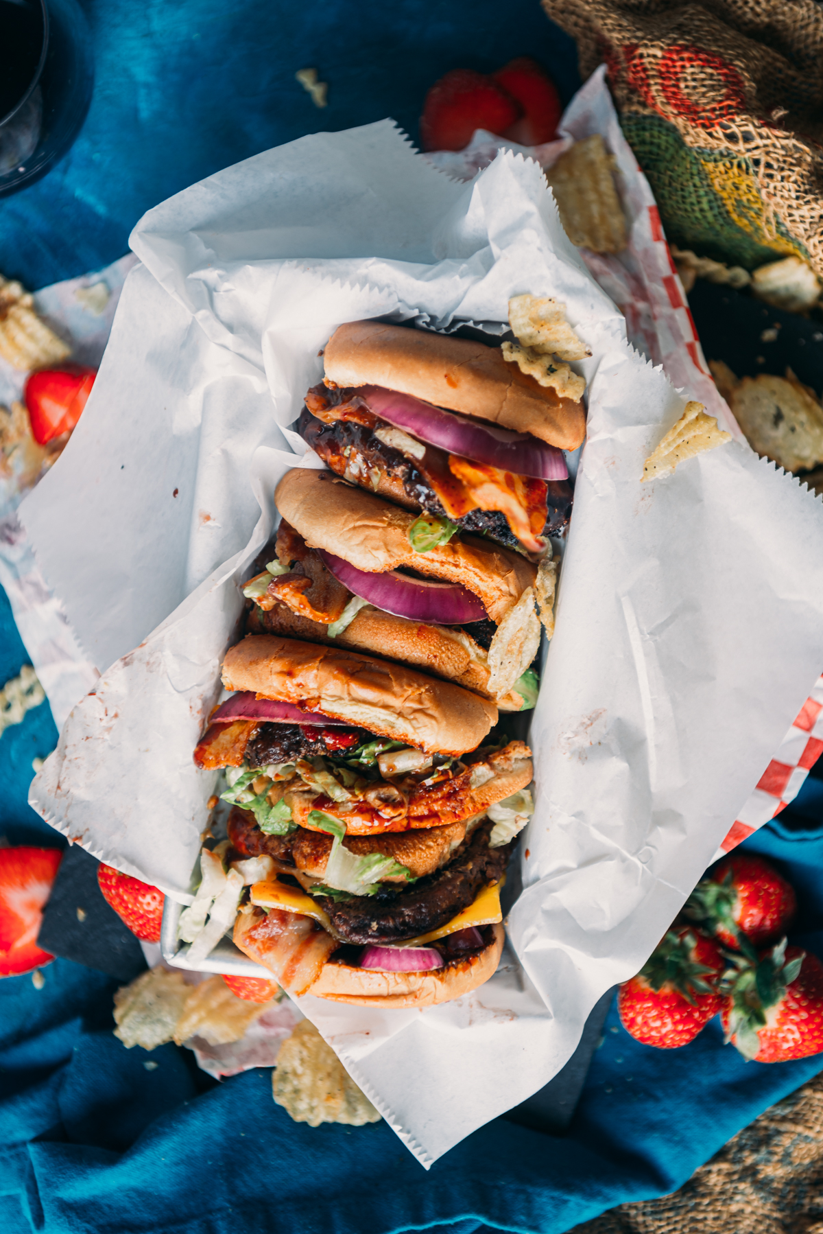 Top view of assorted sandwiches in a paper-lined basket, surrounded by scattered potato chips and whole strawberries on a blue fabric surface.