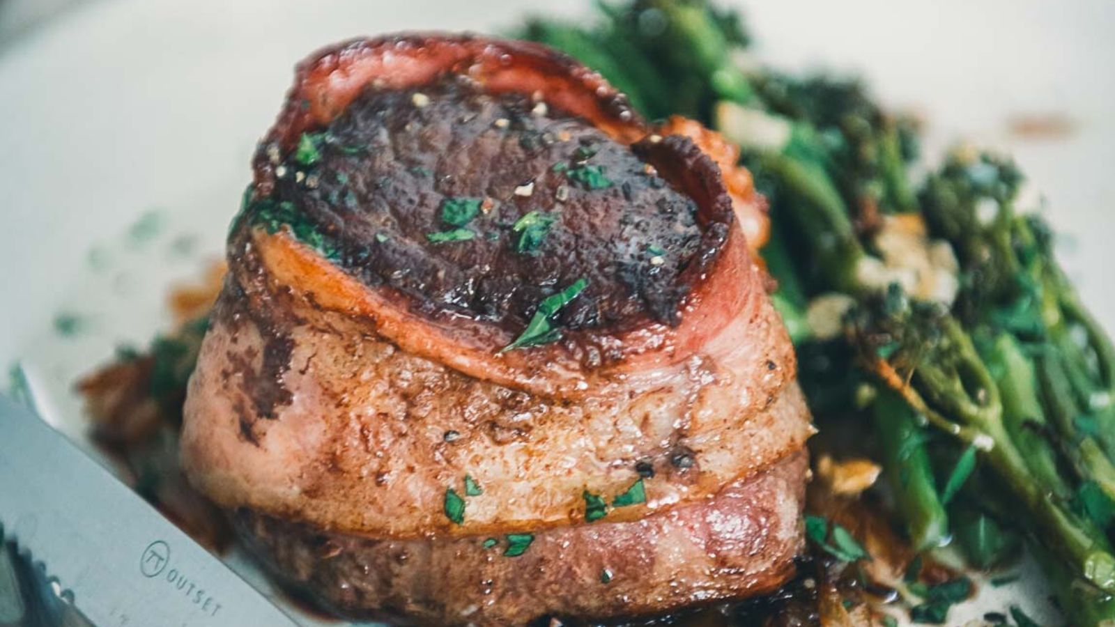 A bacon-wrapped steak served on a plate with a side of sautéed green vegetables. A knife is placed beside the steak.