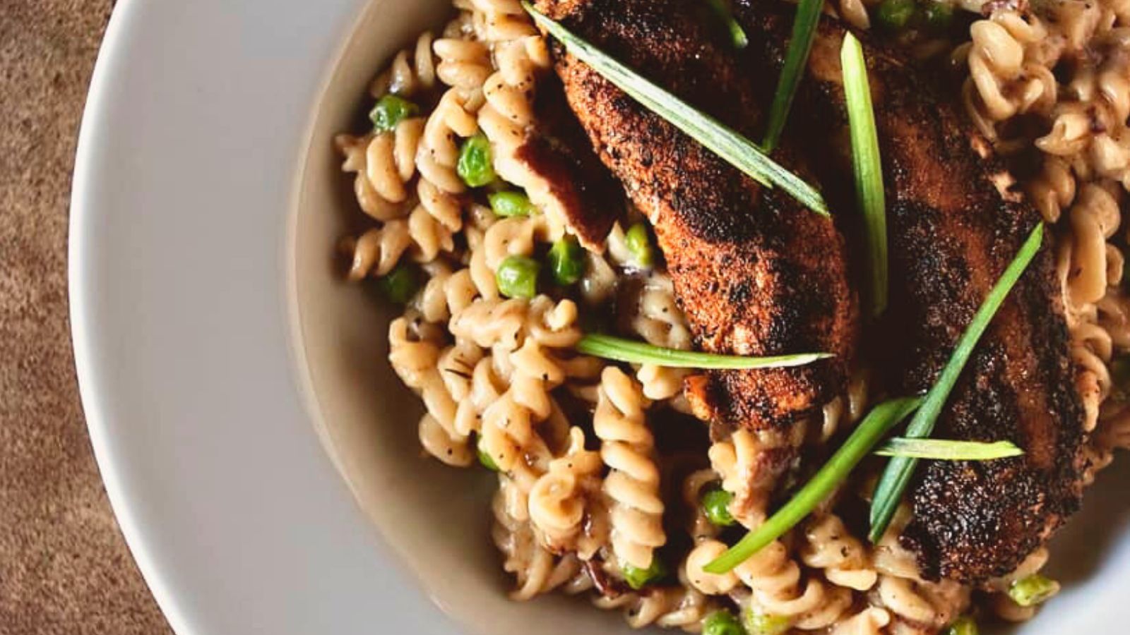 A plate of spiral pasta with peas, topped with grilled meat and garnished with green herbs.