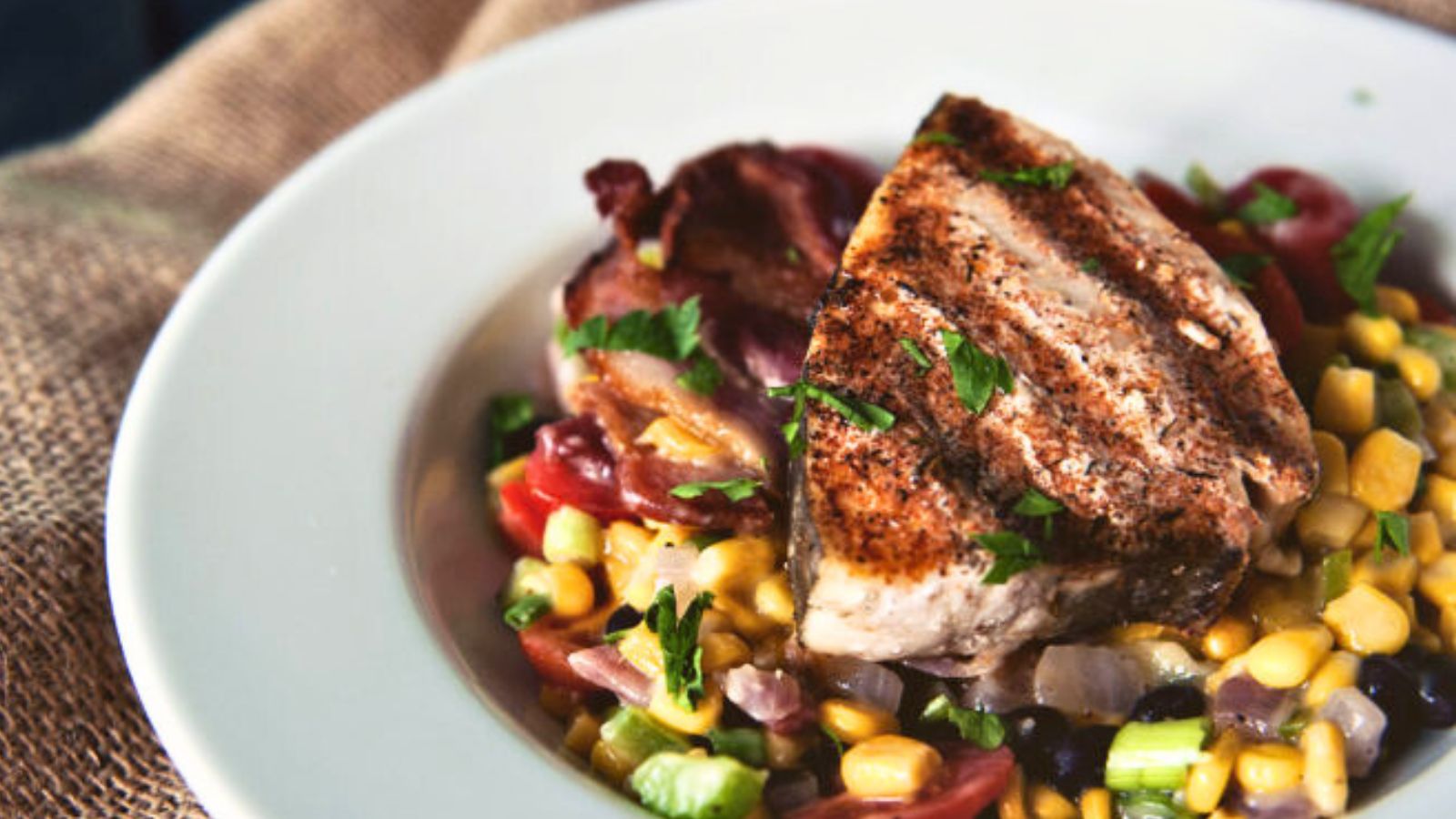 Grilled chicken breast on a bed of mixed vegetables including corn, tomatoes, and celery on a white plate, garnished with chopped herbs.