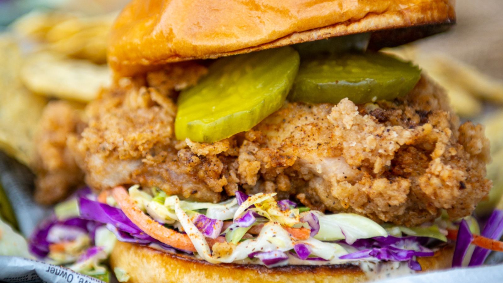 Close-up of a fried chicken sandwich with coleslaw, pickles, and a brioche bun. Chips are visible in the background.