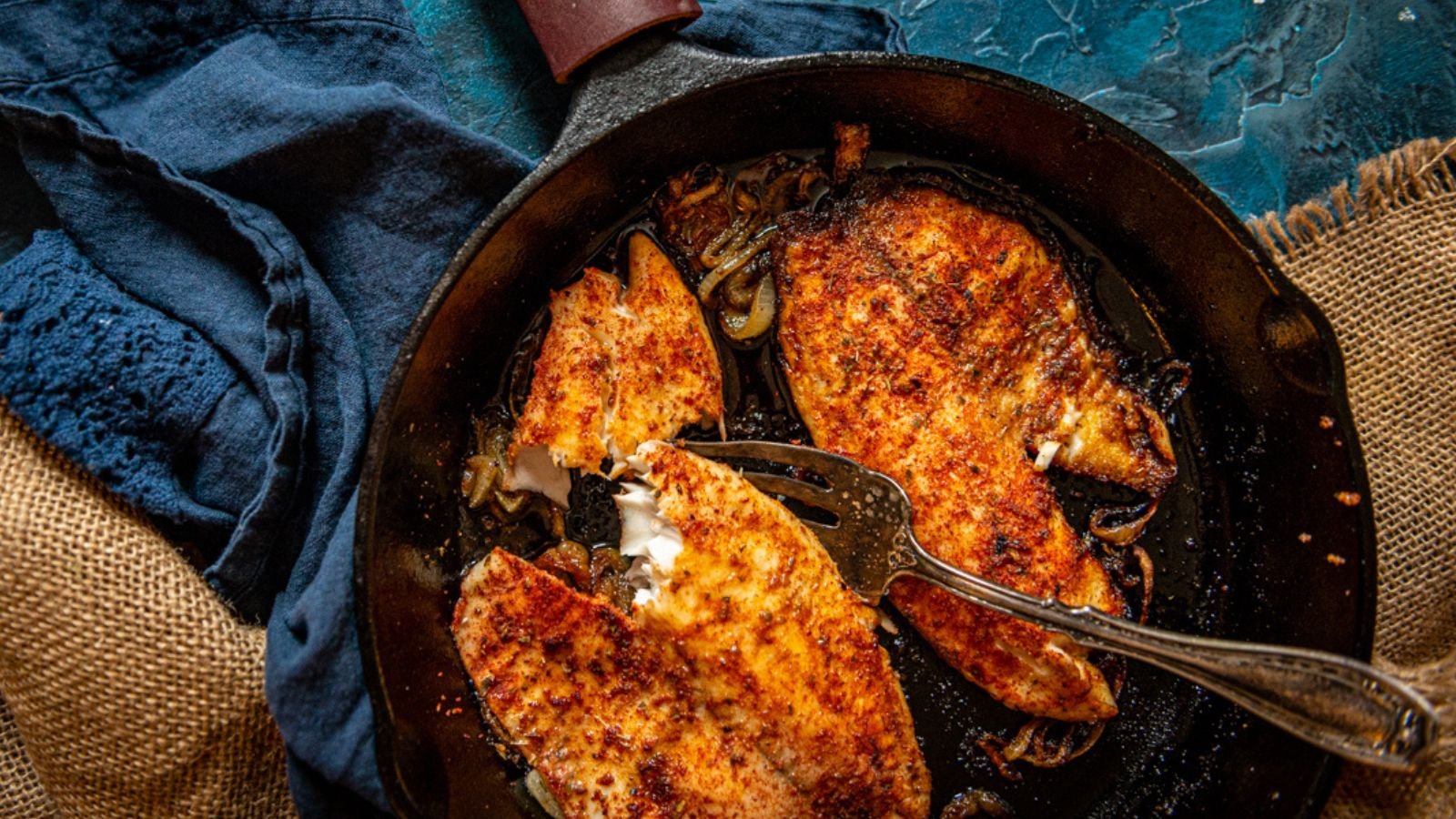 Three seasoned fish fillets sizzle in a skillet, reminiscent of elegant surf and turf recipes perfect for Valentine's Day. A fork gently tears a piece from one fillet, while a cloth lies neatly on the side.