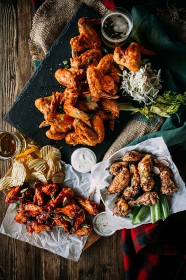 Assorted chicken wings displayed with sauces, celery, coleslaw, potato chips, and beer on a wooden table.