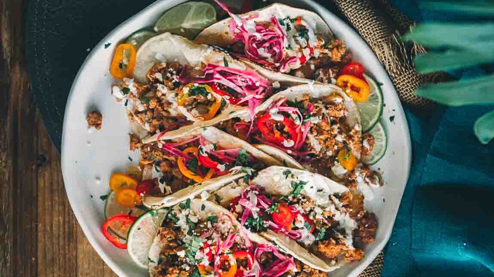 A plate of tacos filled with ground meat, sliced red onions, bell peppers, lime slices, and garnished with herbs and cheese, placed on a wooden table.