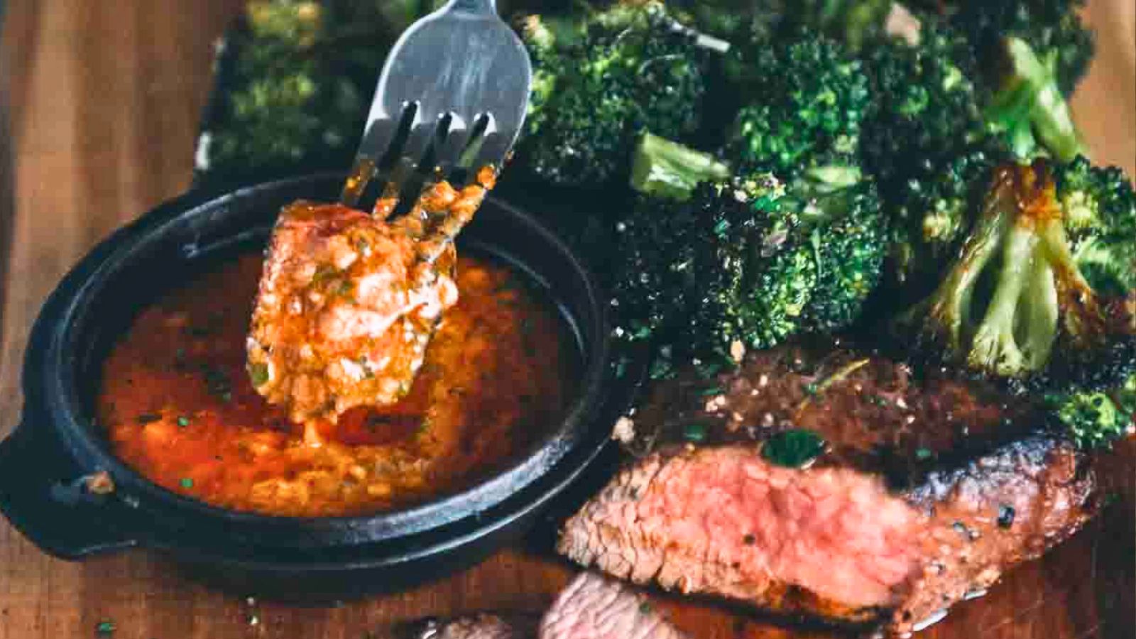 Grilled steak served with broccoli. A piece of steak is being dipped into a red sauce in a small dish.