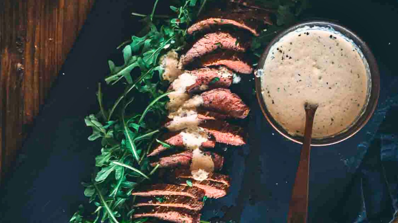 Sliced steak on a dark surface with fresh arugula, accompanied by a bowl of creamy sauce with a spoon.