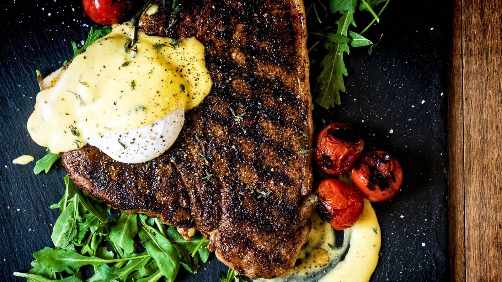 Grilled steak topped with a poached egg and sauce, garnished with arugula and roasted cherry tomatoes, creates a perfect surf and turf twist for Valentine's Day on a sleek black surface.