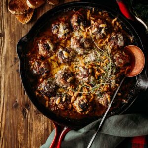 A cast iron skillet with baked french onion meatballs topped with melted cheese and herbs.