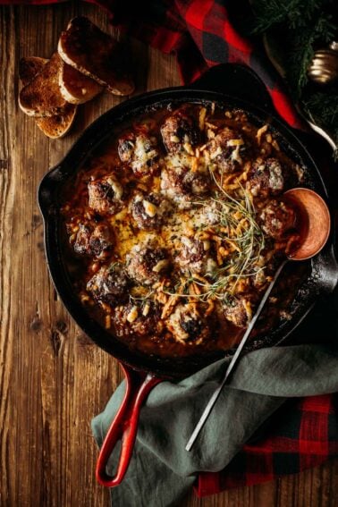 A cast iron skillet with baked french onion meatballs topped with melted cheese and herbs.