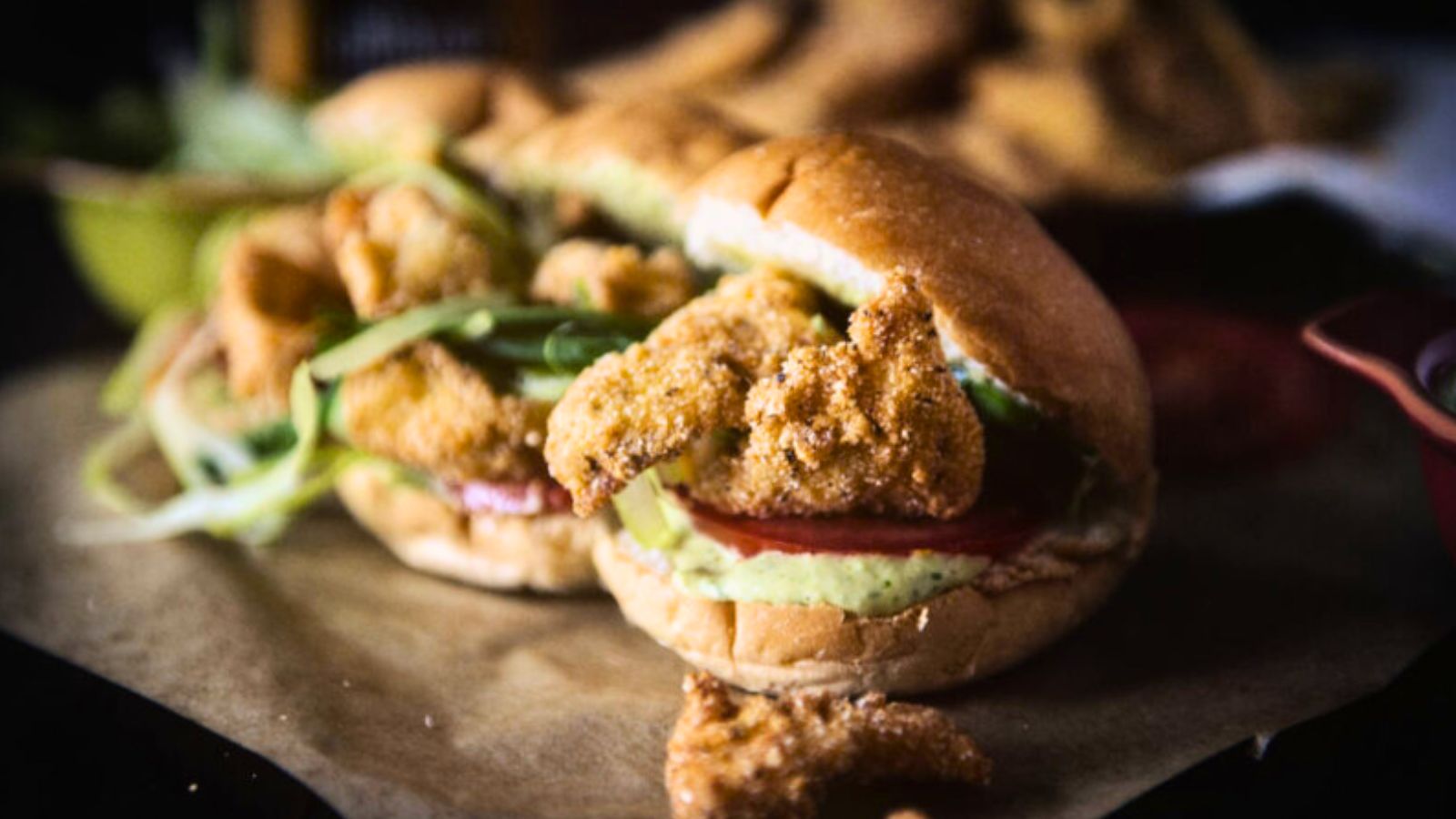 Fried chicken sandwich with lettuce, tomato, and sauce on a bun, placed on parchment paper.