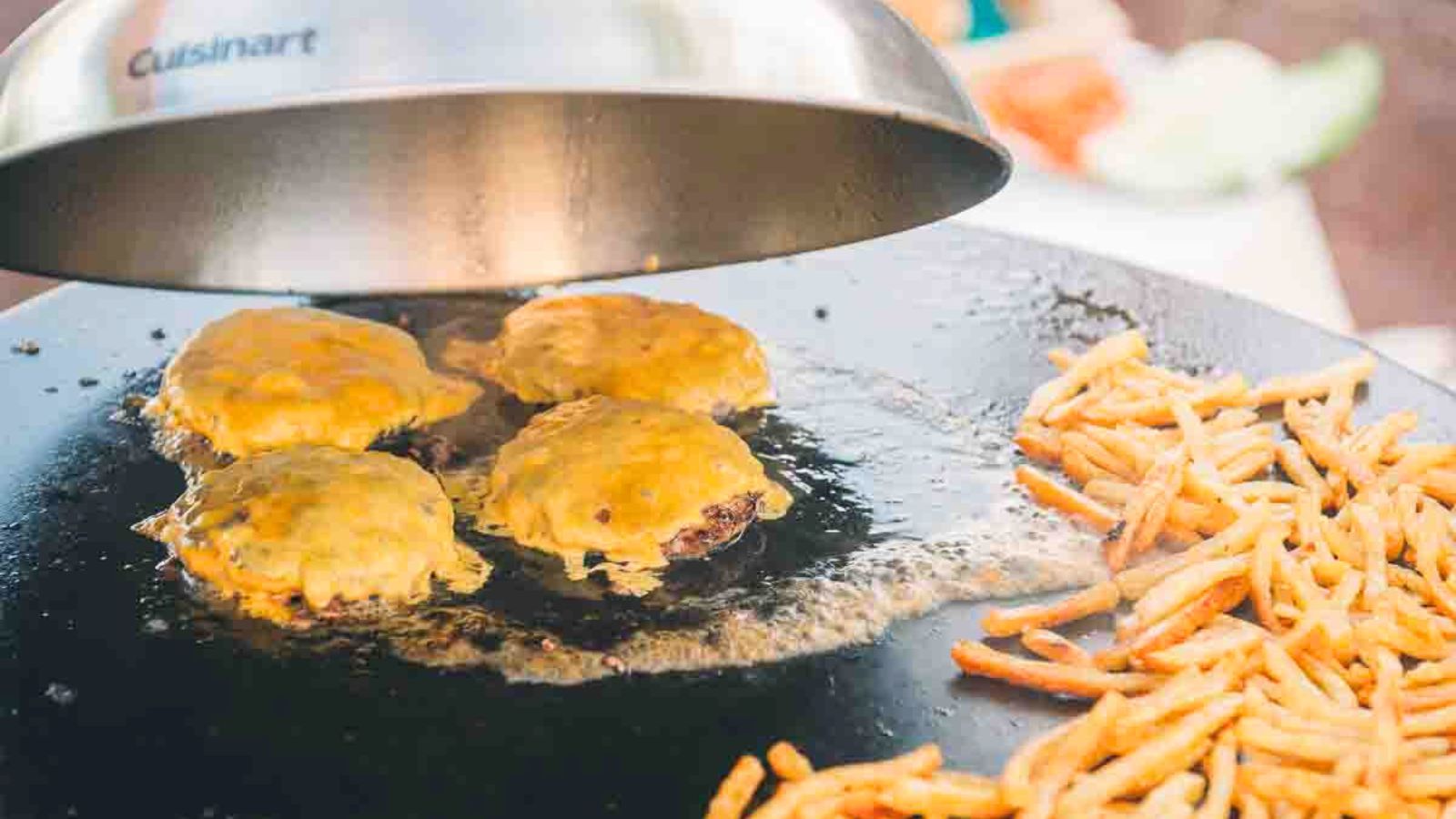 Cheeseburgers cooking under a metal dome on a griddle next to a pile of fries.