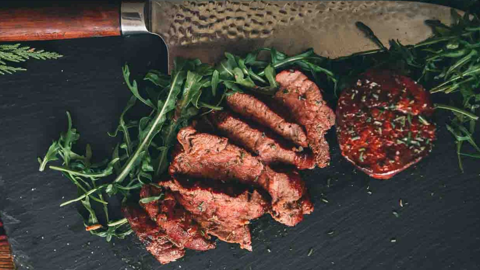 Sliced grilled steak on a black slate with a tomato slice and arugula, served next to a large knife.