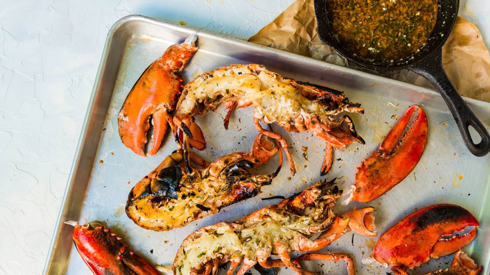 Grilled lobster halves and claws rest on a metal tray, complemented by a skillet of dipping sauce nearby—perfect for elevating your surf and turf recipes for Valentine's Day.