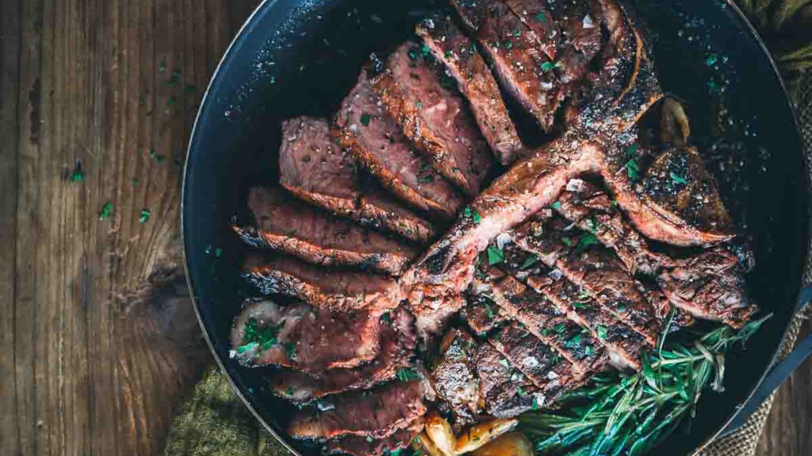 Sliced cooked steak in a black pan, perfect for surf and turf recipes, garnished with herbs on a wooden table background.