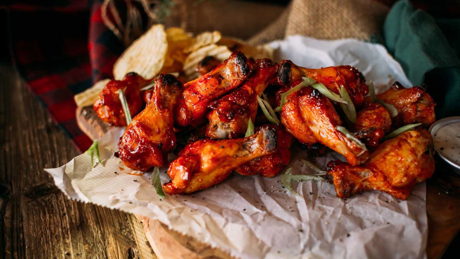 A pile of glazed honey sriracha wings on parchment paper, garnished with green onions, next to potato chips and a small bowl of dipping sauce.