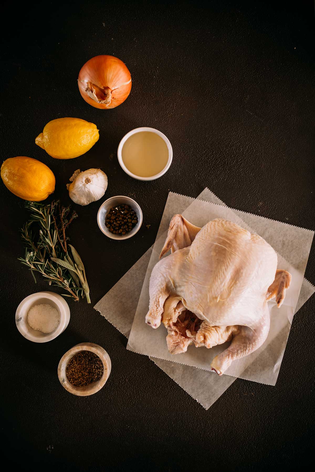 Raw whole chicken on parchment paper with ingredients: two lemons, two onions, garlic, herbs, black pepper, salt, and a cup of oil, arranged on a dark surface.